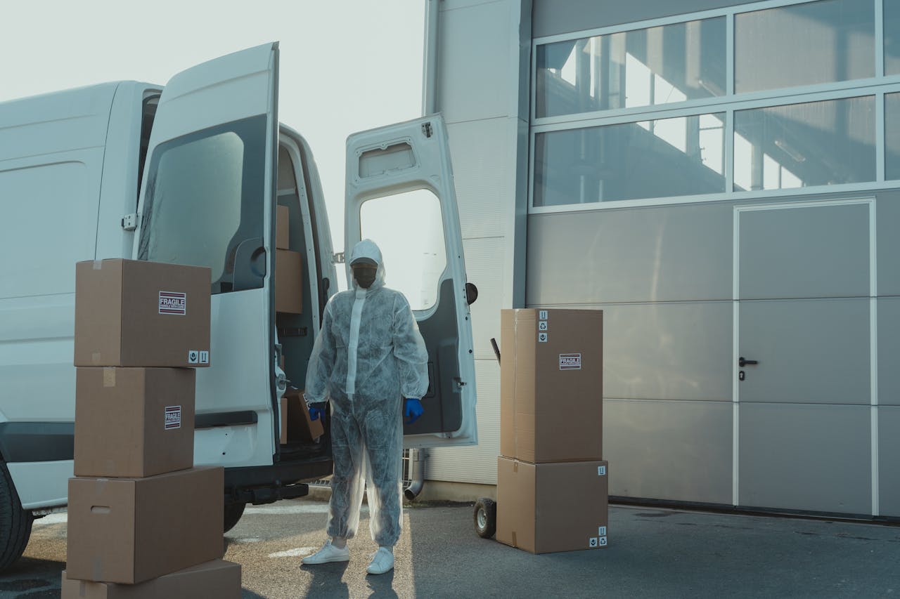 A delivery worker in PPE handles cardboard boxes from a van at an industrial site.