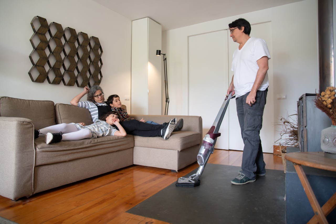 A family relaxes on a sofa while a man vacuums the living room floor, capturing a moment of daily life.